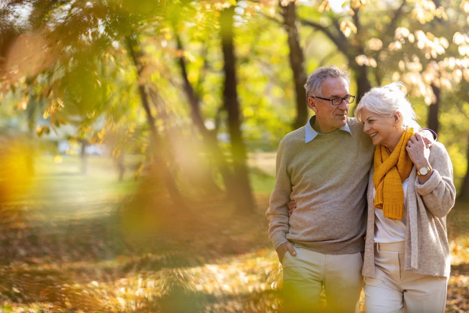 Two older adults walking together through a park with autumn trees and fallen leaves / Deux aînés marchant ensemble dans un parc entouré d’arbres aux couleurs d’automne et de feuilles tombées