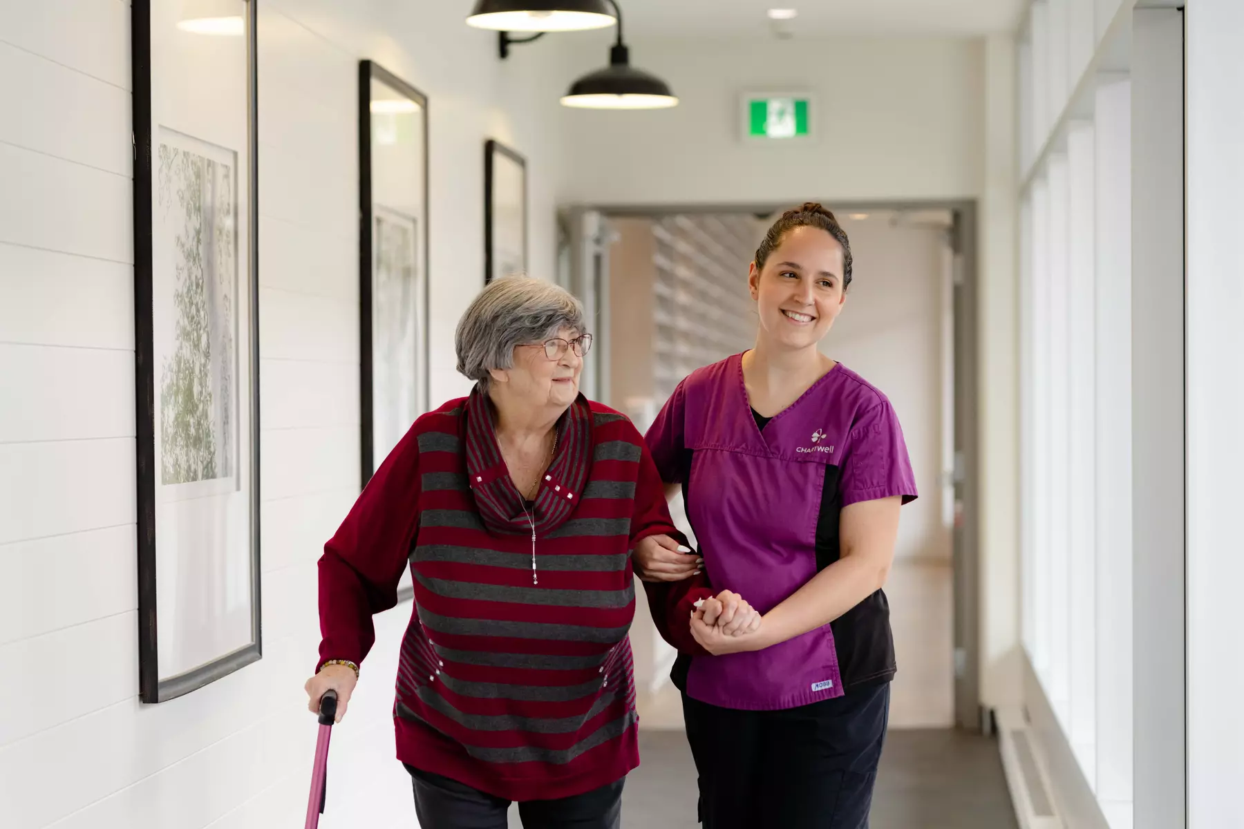 Chartwell staff is helping a residence walking down the hallway with her walking sticking / Le personnel de Chartwell aide une résidence à marcher dans le couloir avec sa canne