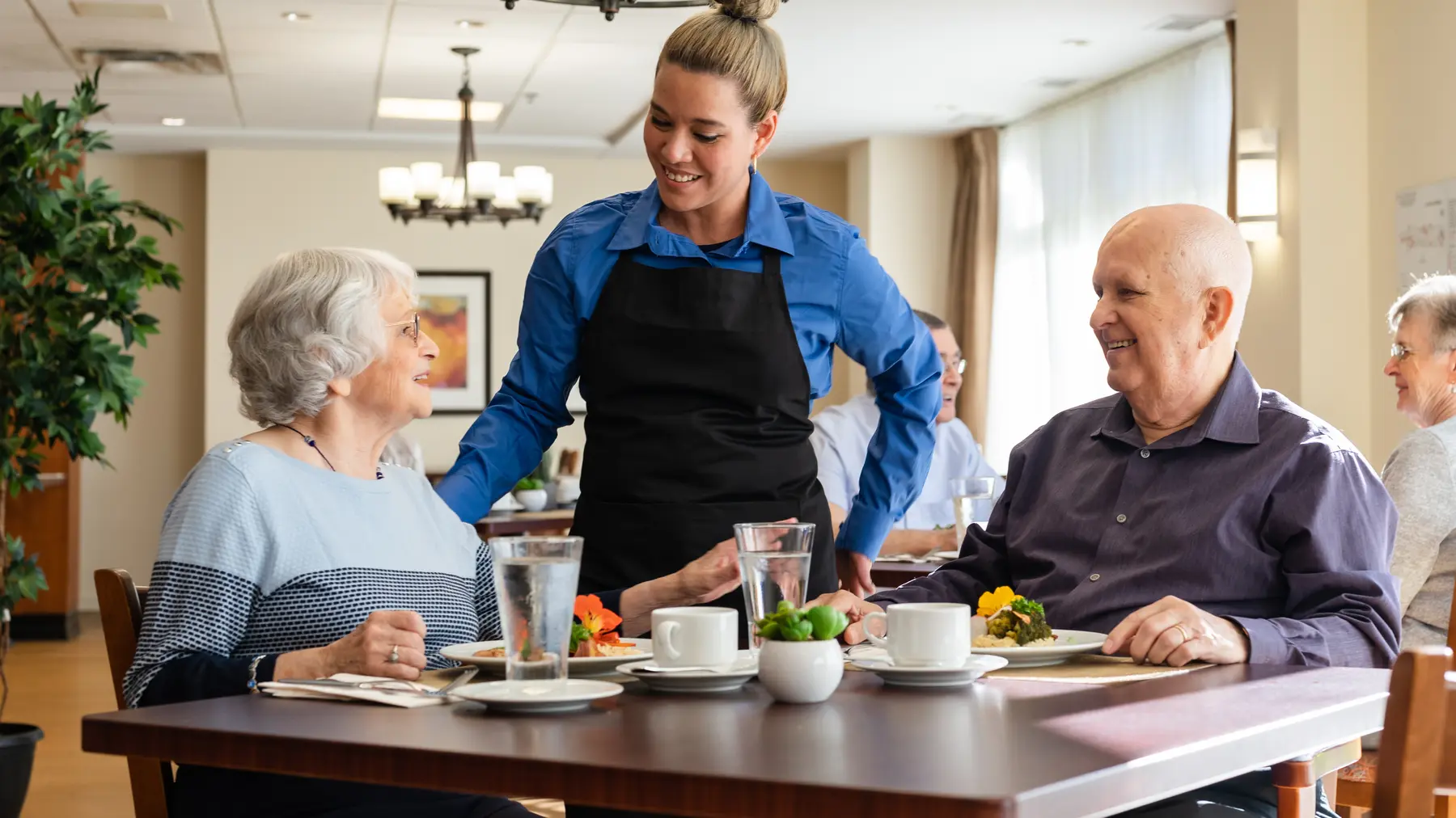 Chartwell Server having a pleasant conversation with the Senior couple at the dinning table / Le serveur de Chartwell a une conversation agréable avec le couple de personnes âgées à la table à manger